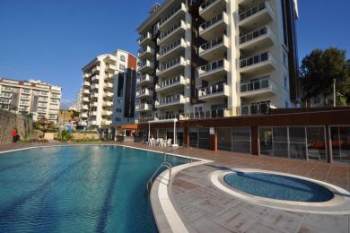 Alanya mulki: Zamonaviy ko'p qavatli uylar orasida quyosh stullari va palma daraxtlari bo'lgan ochiq basseyn.