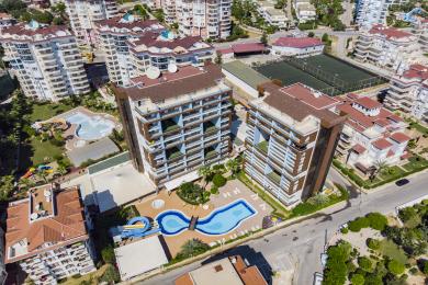 Yorug' Alanya mehmonxonasi krem ​​divan, kulrang kreslolar, yog'och mebellar va ochiq oshxonaga ega.