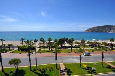 Alanya mulkidagi balkonning quyoshli kunda plyaj, palma daraxtlari va tiniq moviy osmonni namoyish etuvchi manzarasi.
