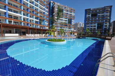 Alanya mulkidagi balkonning yorqin quyosh nurida shahar, basseyn va dengizni namoyish etuvchi ko'rinishi, xotirjam muhit yaratadi.