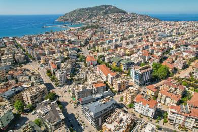 Balkon nekretnine u Alanji s namještajem od pruća, stolom s pločicama, staklenim ogradama i svijetlim, sunčanim pogledom na grad.
