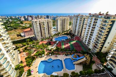Alanya mulkidagi balkon manzarasi basseynlar, tennis kortlari va yorqin quyosh ostida serhosil landshaftni namoyish etadi, bu kurort muhitini yaratadi.