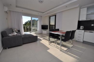 Bright Alanya apartment living area featuring a gray sofa, dining table, and modern white kitchen illuminated by natural light.