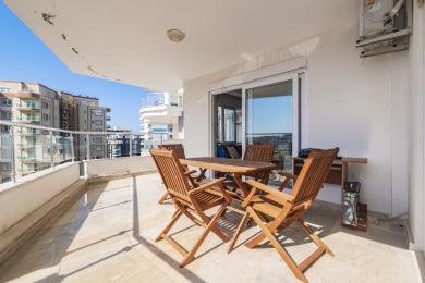 Alanya mulkidagi yog'och stol va stullar, marmar pol, yorqin quyosh nuri va shahar manzarasi bilan jihozlangan balkon.