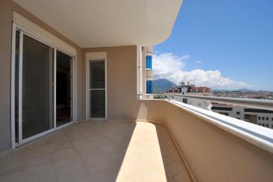 Ochiq rangli Alanya balkonida bej rangli kafel qoplamasi, surma oynali eshiklar va shahar panoramasi manzarasi mavjud.