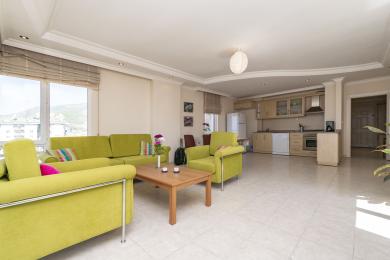 Svetla dnevna soba apartmana u Alanji sa otvorenom kuhinjom, podovima od pločica i udobnim, modernim zelenim sedištima.