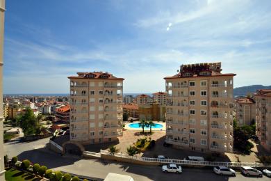Alanya mulkining ko'rinishi, unda ko'p qavatli uylar, basseyn va yorqin, quyoshli osmon ostidagi shahar silueti namoyish etilgan.