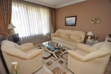 Alanya mulkidagi krem ​​divan, kreslolar, shisha stol va yumshoq tabiiy yorug'lik bilan jihozlangan yashash xonasi.