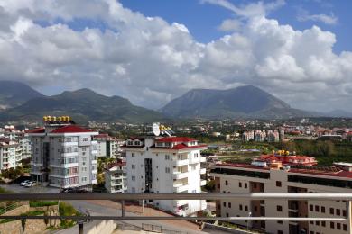 Alanya mulkining balkonidan shahar binolari va uzoqdagi tog'lar qisman bulutli osmon ostida ko'rinadi.