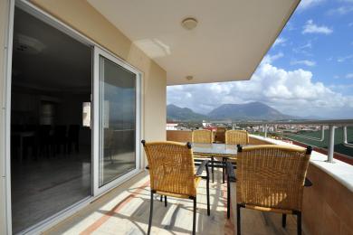 Balkon nemovitosti v Alanyi s proutěným nábytkem a úžasným výhledem na hory, vytvářející relaxační venkovní obytný prostor.