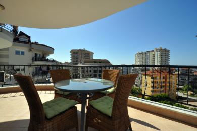 Alanya mulkidagi balkon, to‘qilgan stullar, shisha stol va yorqin, quyoshli osmon ostidagi shahar manzarasi.