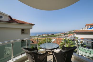 Balkon nekretnine u Alanji sa pletenim namještajem i staklenom ogradom, nudi zadivljujući pogled na more i grad u jarkoj sunčevoj svjetlosti.