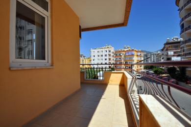 Quyoshli Alanya balkonida kafel qoplamasi, to'q sariq devorlar va tiniq moviy osmon ostida shahar manzarasi.