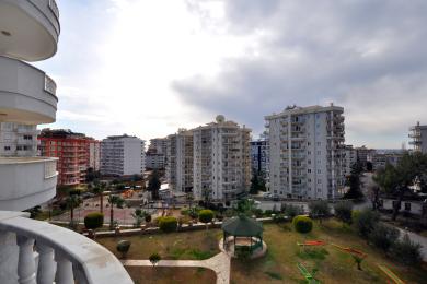 Alanya mulkidagi balkonning obodonlashtirilgan bog', yaqin atrofdagi binolar va bulutli osmonga qaragan manzarasi.