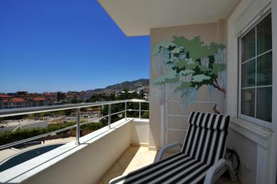 Балкон у нерухомасці ў Аланіі з відам на горад, размаляванай фрэскай і паласатым шэзлонгам у яркім сонечным святле.