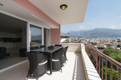 Alanya mulkidagi balkon, toʻqilgan ovqatlanish mebellari, kafel pol va shahar va togʻning panoramik manzaralari bilan.