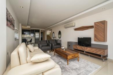 Yorug' Alanya mehmonxonasi krem ​​divan, kulrang kreslolar, yog'och mebellar va ochiq oshxonaga ega.