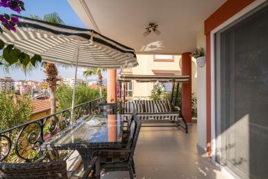 Alanya-Balkon mit Korbmöbeln, gestreifter Schaukel und Stadtblick, der einen entspannenden Wohnbereich im Freien schafft.