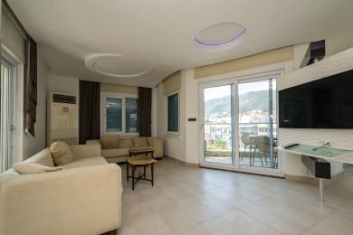 Alanya mulkidagi bej divanlar, kafel pol, balkon va zamonaviy yoritishga ega yashash xonasi.