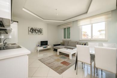 Svetla dnevna soba u apartmanu u Alanji sadrži sivi ugaoni kauč, beli trpezarijski set i podne pločice.