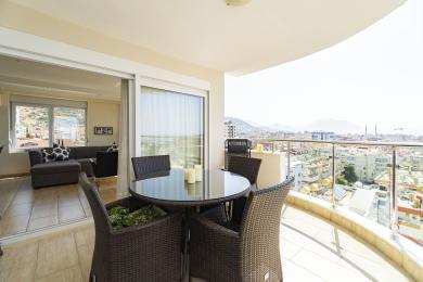 Balkon nekretnine u Alanji sa nameštajem od pruća, staklenim stolom i panoramskim pogledom na grad, okupan jarkim suncem.