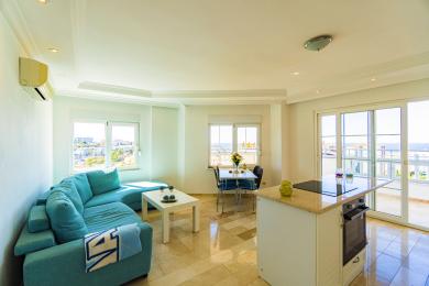 Zonă de locuit luminoasă într-un apartament din Alanya, cu canapea turcoaz, podele de marmură și acces la balcon cu vedere la mare.