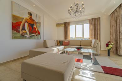 Yorqin Alanya mehmonxonasi katta bej rangli divan, marmar pollar va dekorativ qandilga ega.