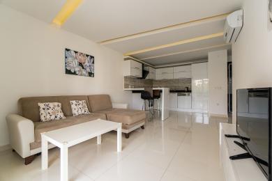 Alanja apartman dnevna soba sa modernom belom kuhinjom, bež sofom i svetlim, popločanim podovima koji stvaraju čistu, prozračnu atmosferu.