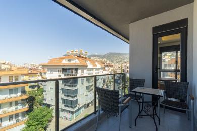 Balkon nekretnine u Alanji s pogledom na grad, staklenom ogradom, podom od pločica i vanjskim sjedećom garniturom pod jarkim suncem.