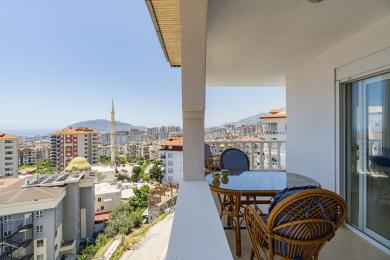 Balkon nekretnine u Alanji sa namještajem od ratana i staklenim stolom, koji nudi svijetli pogled na grad i more.