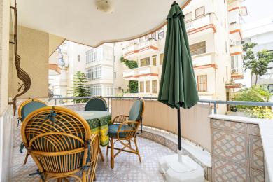 Balkon nekretnine u Alanji sa nameštajem od pruća, podom od šarenih pločica i zelenim suncobranom za život na otvorenom u hladu.
