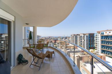 Alanya mulkidagi balkon, toʻqilgan mebellar, kafel qoplamasi va yorqin, quyoshli osmon ostida shahar panoramasi.