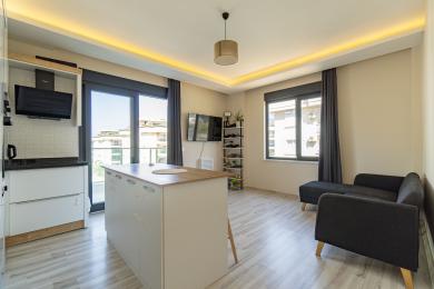 Yorqin Alanya kvartirasi, orol oshxonasi, balkon chiqishi va kulrang divanli zamonaviy yashash maydoniga ega.