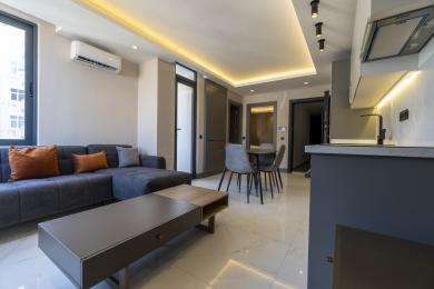 Deze woning in Alanya beschikt over een moderne woonkamer met een strakke grijze bank, een eethoek en heldere, inbouwverlichting, wat een gezellige sfeer creëert.