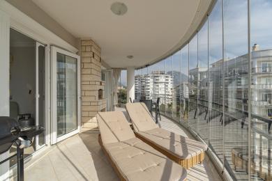 Balkón nemovitosti v Alanyi se skleněnými stěnami, dlaždicovou podlahou, lehátky a výhledem na město, vytvářející světlou a relaxační atmosféru.