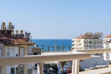 Μπαλκόνι ακινήτου στην Αλάνια με θέα στη Μεσόγειο Θάλασσα, φοίνικες και καθαρό γαλάζιο ουρανό.