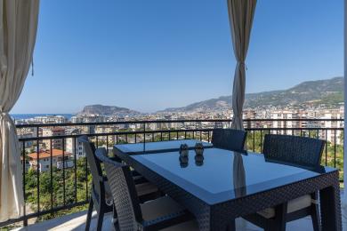Alanya-Immobilienbalkon mit Korbmöbeln, Glastisch und atemberaubendem Blick auf die Stadt und das Meer.