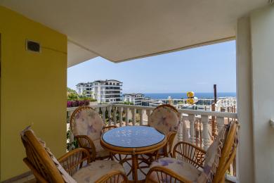 "Alanya mulkidagi balkon, to'qilgan mebellar, gulli yostiqlar va yorqin kunduzgi yorug'likda ajoyib dengiz manzarasi bilan."