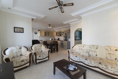 Alanya property living room featuring floral sofas, tile flooring, and an open-plan kitchen, creating a bright and airy atmosphere.
