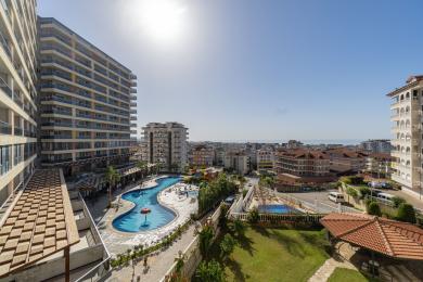 Pogled na nekretninu u Alanji: Balkon s pogledom na veliki bazen, gradsku liniju i Sredozemno more po sunčanom danu.