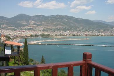 Balkon nekretnine u Alanji s drvenom ogradom s pogledom na tirkizno more i panoramu grada, okupan jarkim suncem.