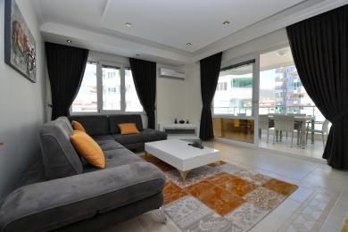 Yorug' Alanya mehmonxonasi kulrang divan, balkon, qora pardalar va zamonaviy oq mebellarga ega.