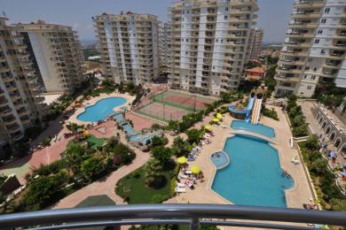 Alanya woning balkon uitzicht toont zwembaden, tennisbanen en weelderig groen onder helder zonlicht, waardoor een resort sfeer ontstaat.