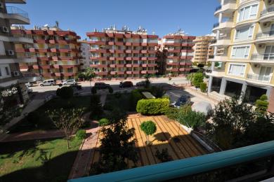 Alanya ingatlan erkélyéről kilátás egy parkosított udvarra és a közeli apartmanházakra egy napsütéses napon.